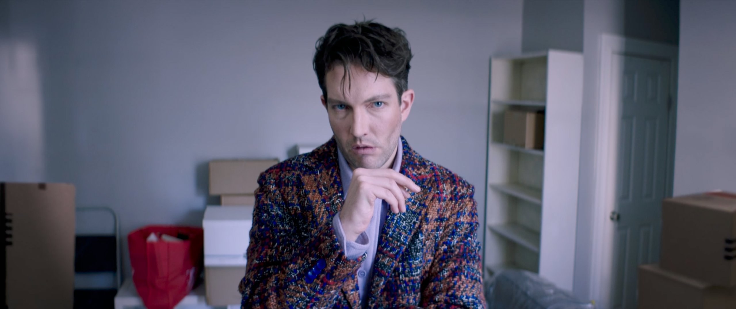 Man in patterned blazer, hand at chin. Boxes stacked, empty shelves behind.