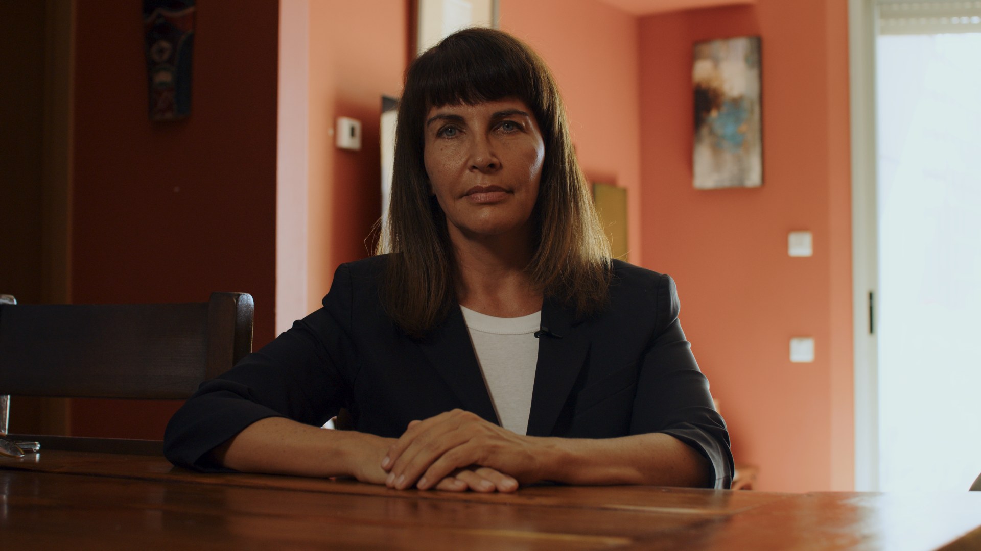Interview at a wooden table. Coral walls, hands folded.