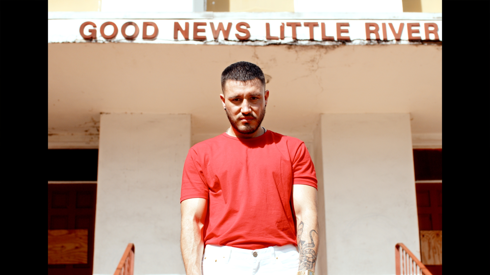 Standing under the Good News Little River sign. Red tee, daylight.